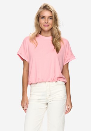 Mujer rubia con cabello suelto y ondulado, vestida con una blusa rosa de manga corta y pantalones blancos de talle alto, de pie frente a un fondo liso.