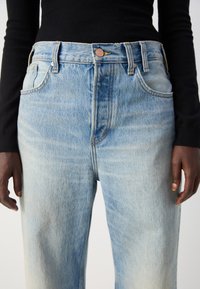 Jean en denim bleu clair avec une taille haute, une coupe décontractée et une finition délavée. Comprend des poches avant et une fermeture à bouton en métal. Surface texturée.