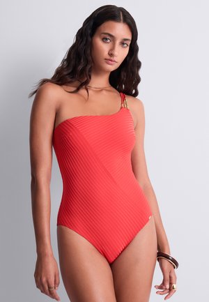 Femme aux longs cheveux ondulés et foncés portant un maillot de bain rouge une épaule côtelé avec des accents dorés, tenant des bracelets à son poignet droit.