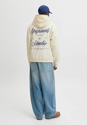 Person trägt cremefarbenen Hoodie mit blauem "Originals Studio" Text auf dem Rücken, weite hellblaue Jeans, braune Schuhe und eine blaue Baseballkappe, rückwärts gewandt.