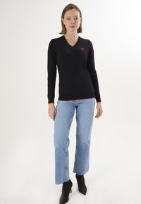 Schwarzer, langärmliger V-Ausschnitt-Pullover mit einem kleinen roten Herzemblem, kombiniert mit hellblauen, weiten Jeans und schwarzen Knöchelstiefeln.