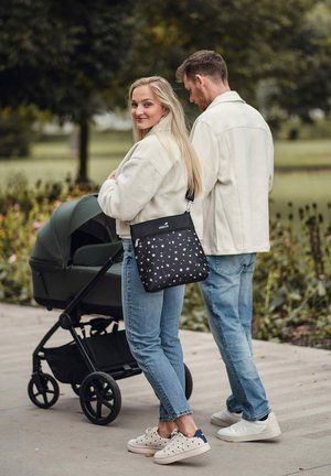 Černá taška přes rameno s květinovým vzorem a zipem, nošená přes tělo, vedle tmavě zeleného kočárku na cestě v parku.