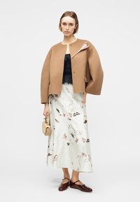 Femme portant une veste surdimensionnée beige avec une broche fleurie, un haut noir, une jupe blanche à motifs floraux, des ballerines bordeaux, tenant un petit sac à main beige.