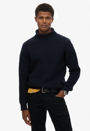 Marineblauwe gebreide coltruik met een textuurpatroon, gecombineerd met een gele onderhemd, zwarte broek en een zwarte riem.