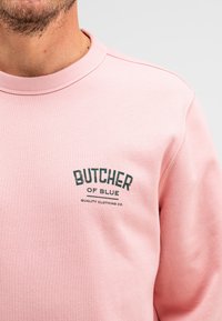 Roze sweatshirt met ribbelkraag en -manchetten, met een zwart logo met de tekst "BUTCHER OF BLUE" en "QUALITY CLOTHING CO." op de borst.