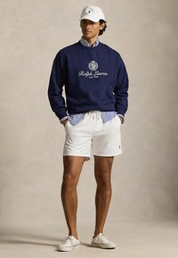 Mladý muž ve tmavě modré mikině Ralph Lauren, pruhované košili, bílých šortkách, bílé čepici a bílých teniskách, stojí s rukama v kapsách.