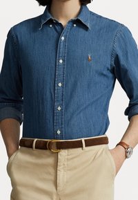 Camisa de mezclilla azul, con cuello abotonado y logo bordado. Combinada con pantalones beige, cinturón marrón y un reloj con correa de cuero.