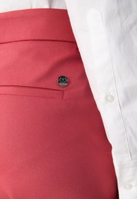 Gros plan sur un pantalon rouge avec un petit logo de marque métallique près d'une poche arrière, porté avec une chemise blanche boutonnée.