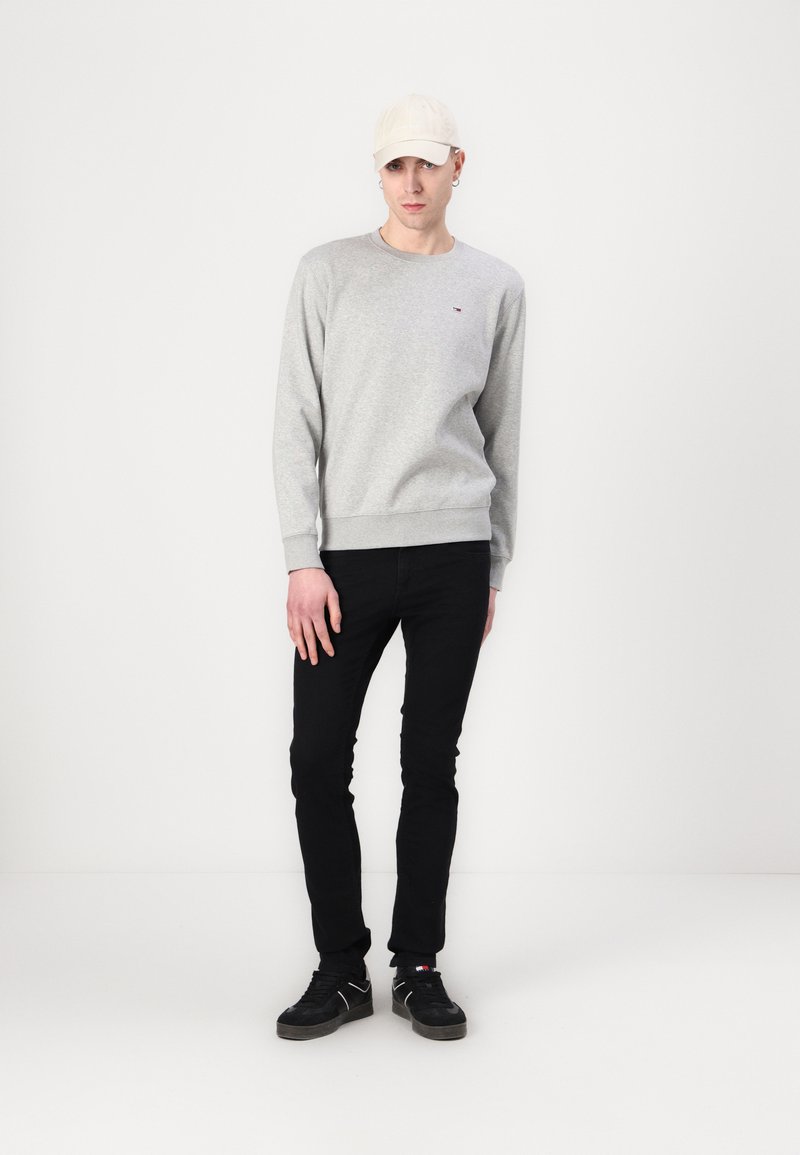 Szary sweter z ściągaczami, zestawiony z czarnymi dopasowanymi dżinsami i czarnymi sneakersami. Jasny kapelusz dopełnia całość.