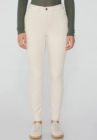 Vaqueros ajustados beige claro hechos de una tela elástica, con una cintura de altura media, bolsillos frontales y una textura suave. Llevados con zapatillas ligeras.