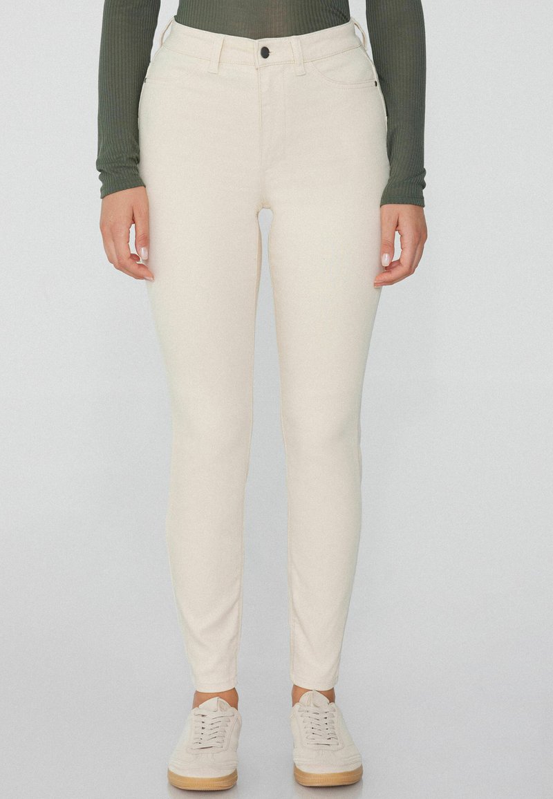 Vaqueros ajustados beige claro hechos de una tela elástica, con una cintura de altura media, bolsillos frontales y una textura suave. Llevados con zapatillas ligeras.
