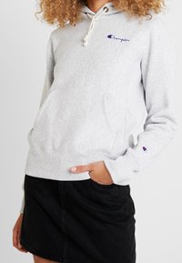 Grauer Hoodie aus weichem Material, mit einer Kängurutasche, verstellbarem Kapuzenzug und besticktem Logo auf der linken Brust. Darunter ein schwarzer Rock.