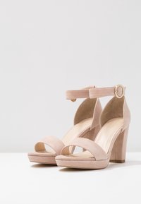 Pár béžových semišových sandálů na vysokém podpatku s řemínky kolem kotníků a zlatou sponou, umístěných na bílém povrchu proti jednolitému pozadí.