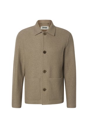 Jeune homme debout portant une veste marron boutonnée, un pantalon en velours côtelé marron foncé et des bottes noires sur un fond gris clair uniforme.