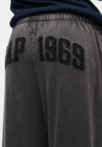 Κοντινό πλάνο γκρι παντελονιού με ελαστική ζώνη μέσης που φέρει μαύρο κεντητό κείμενο «GAP 1969» στο πίσω μέρος.