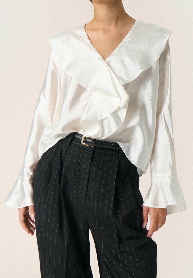 Femme portant un chemisier en satin blanc avec col et poignets à volants, associé à un pantalon taille haute à rayures noires et une ceinture noire.