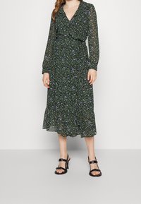 Mujer vistiendo un vestido midi negro de mangas largas con un pequeño estampado de flores azules y verdes, y sandalias negras de tacón con tiras en los tobillos.