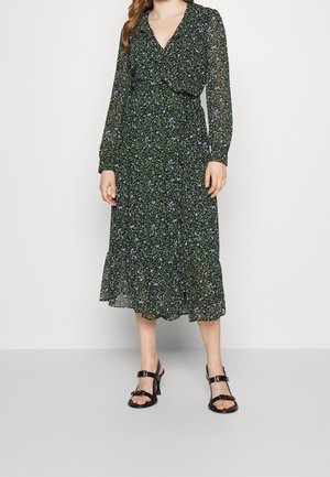 Femme portant une robe midi noire à manches longues avec un petit motif floral bleu et vert et des sandales à talons noires avec des brides à la cheville.