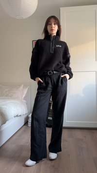 Μαύρο φλις πουλόβερ με ψηλό γιακά και λογότυπο GAP· σετ με φαρδιά μαύρα παντελόνια. Παπούτσια λευκά slip-on. Φόντο δάπεδο από ξύλο.