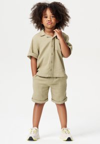 Chemise vert clair texturée à manches courtes avec une poche poitrine, accompagnée de shorts assortis. Baskets blanches avec des accents jaunes.