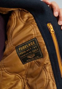 Černý vlněný kabát s vnitřní zlatou prošívanou podšívkou, vybavený štítkem s textem "FUSELAGE" a žlutým zipem na boku.