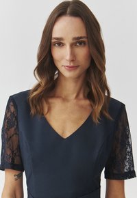 Marineblaues Kleid mit tiefem V-Ausschnitt, kurzen Spitzensäumen und tailliertem Oberteil. Die Textur ist glatt mit zarten Spitzenverzierungen an den Ärmeln.