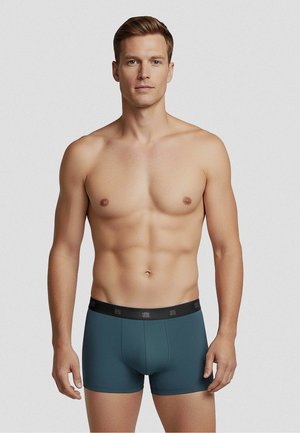 Lihaksikas lyhyttukkainen mies, joka pukeutunut teal-värisiin bokserialushousuihin, musta vyötärönauha, seisoo yksiväristä vaaleanharmaata taustaa vasten.