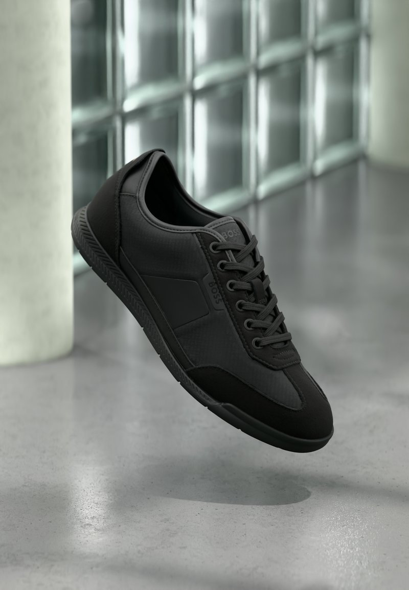 Baskets basses noires à lacets, marquées "BOSS", flottant devant un sol gris et un mur en blocs de verre.