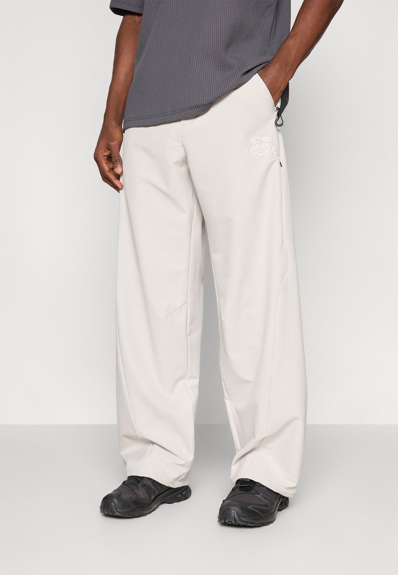 Calças largas bege com uma textura suave, apresentando um bolso lateral e um detalhe em logotipo branco. Combinadas com uma camisa cinza escuro e sapatos pretos.