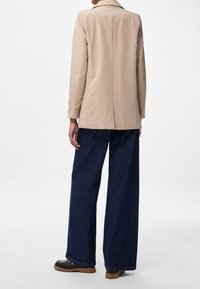 Blazer beige à la coupe structurée, fente unique au dos, associé à un pantalon en denim bleu foncé ample et des chaussures foncées.