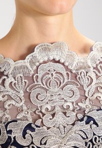 Krajkov dress s detailní stříbrnou květinovou výšivkou na průhledné látce a volánkovým výstřihem. Námořnické akcenty na lemu.