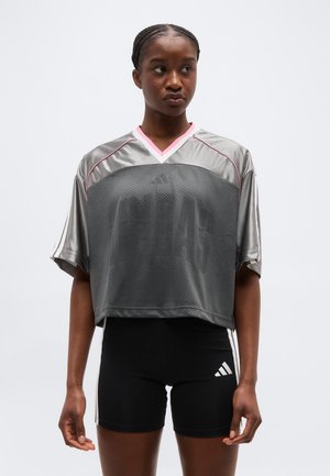 Žena s pletenými vlasmi, ktorá nosí voľný sivý a ružový športový dres a čierne cyklistické šortky Adidas, stojaca na jednoduchom pozadí.