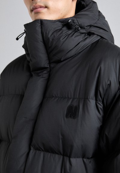 Chaqueta puff negra con cuello alto, acabado brillante, textura acolchada y un sutil logotipo bordado en el lado izquierdo del pecho.