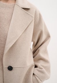 Beige vlněný kabát s výstřihem ve tvaru klopy, jedním černým knoflíkem a spadlými rameny. Měkká textilie s volným střihem a útulným pleteným svetrem pod ním.