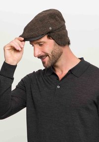 Mies, jolla on parta, pukeutuneena harmaaseen pitkään hihaiseen polo-paitaan ja ruskeaan tweed-malliseen lippalakkiin, säätää lippalakkia oikealla kädellään, hymyilee ja katsoo alaspäin.