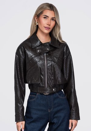 Femme aux longs cheveux blonds portant une veste en cuir noire texturée et un jean bleu foncé, debout devant un fond clair uni.