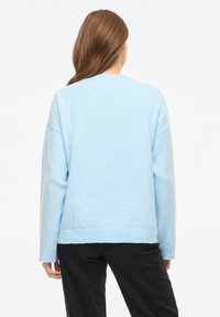Svetlo modra pulover iz mehkega materiala, z ohlapnim krojem, dolgimi rokavi in rebrastim robom. Brez opaznih vzorcev ali kovinskih detajlov.