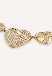 Bracciale in tono oro con maglie a forma di cuore ricoperte di strass trasparenti, incise con "GUESS", che mostrano finiture lisce e texture diverse.