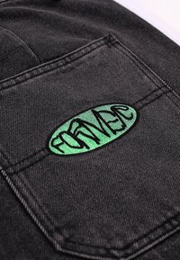 Tela de mezclilla negra con costuras visibles y un parche ovalado verde bordado con la palabra "FORNACE" en hilo negro.