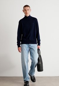 Marinefarbener Rollkragenpullover, lockere Passform, kombiniert mit hellblauen geraden Jeans. Das Model hält eine schwarze Tasche und trägt schwarze Schuhe. Glatte Stoffstruktur.