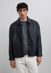 Hombre con una chaqueta de cuero negra sobre un suéter gris y pantalones blancos, de pie contra un fondo gris claro liso.