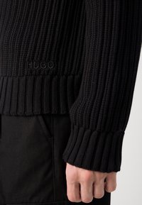 Gros plan sur une manche et un poignet de pull en maille côtelée noire sur un pantalon noir, montrant le logo discret "HUGO" sur le bas de la manche.