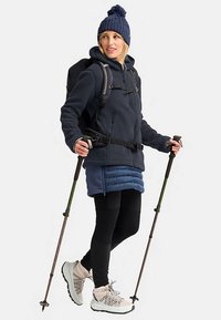 Fleecejack in marineblauw, blauwe gewatteerde rok, zwarte legging en lichte wandelschoenen. Inclusief rugzak en wandelstokken.