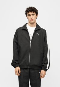 Νεαρός άνδρας που φοράει μαύρο αθλητικό μπουφάν και παντελόνι Puma με λευκές ρίγες, στέκεται μπροστά σε απλό ανοιχτό φόντο.