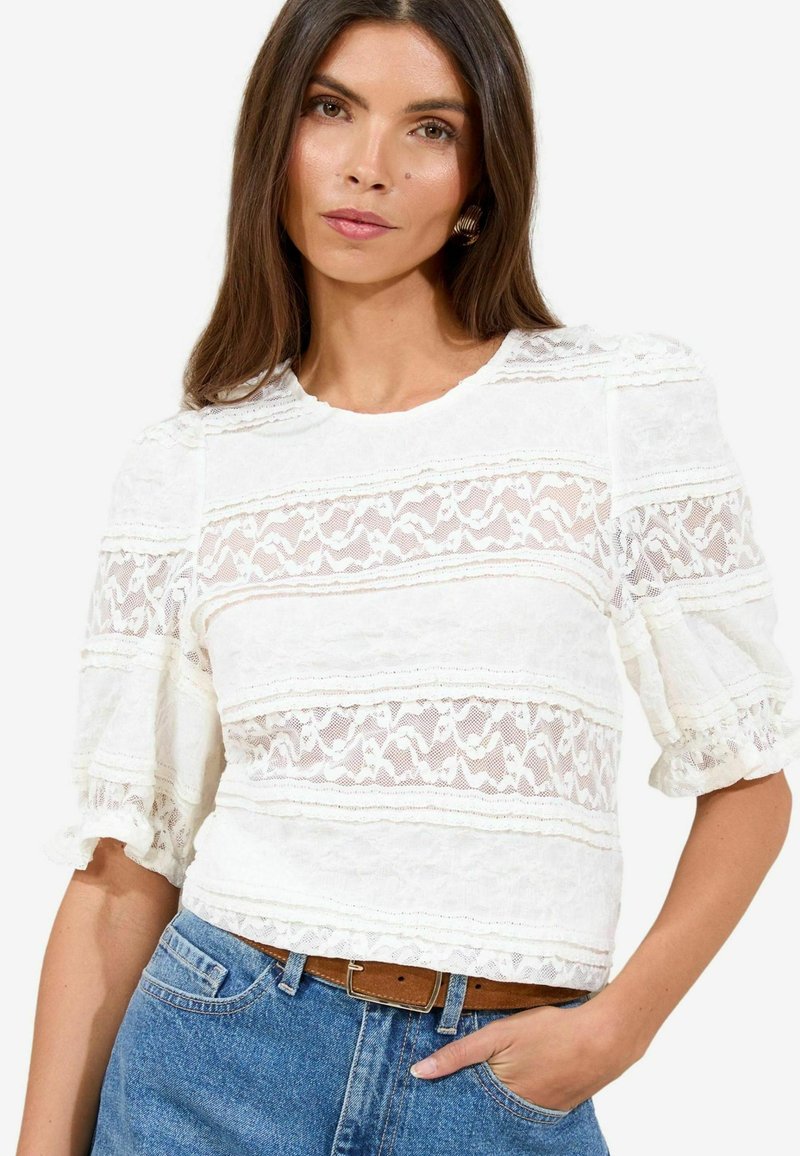 Top in pizzo bianco con maniche a palloncino, caratterizzato da un motivo testurizzato e sezioni in tessuto trasparente. Abbinato a jeans azzurri chiari e una cintura marrone.