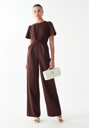 Brun bredbenet jumpsuit med korte pufærmer og et snoet frontdetalje. Sættet er parret med en hvid struktureret håndtaske og lyse sandaler.
