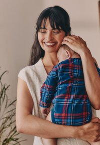 Kobieta o długich ciemnych włosach w białym dzianinowym topie trzyma niemowlę ubrane w niebieską i czerwoną koszulę w kratę. W tle widać rośliny doniczkowe.