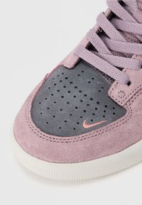 Nærbilde av en rosa og grå semsket sneaker med perforeringer, rosa lisser og en liten rosa Nike swoosh på tåboksen.