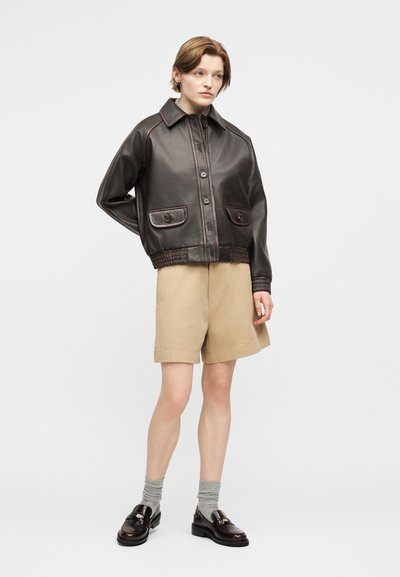 Άτομο ντυμένο με σκούρο καφέ δερμάτινο μπουφάν, μπεζ σορτς μέχρι το γόνατο, γκρι κάλτσες και σκούρα καφέ μοκασίνια, στέκεται μπροστά σε απλό φόντο.