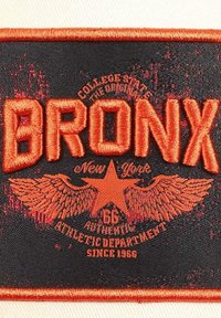 Schwarzer gewebter Patch mit lebhaftem orangefarbenem Stick. Enthält "BRONX" in fetten Buchstaben, ein Adlerdesign, einen Stern und einen Text, der eine Sportabteilung angibt.